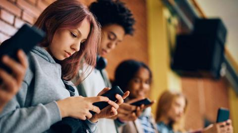 Four school pupils are standing against a brick wall, not talking to each other as they each look down at a phone in their hands