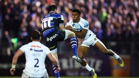 Two rugby players, one in blue, the other in white jumping in the air, competing for rugby ball as crowd watch in background