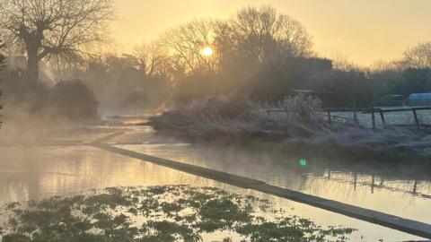 Fog and mist shroud a river and nearby grassland. Sun rising in the distance.  