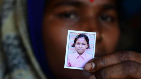 An Indian woman in blurred focus holds out a small photo of her daughter who died of Guillain Barre Syndrome during an outbreak in 2019