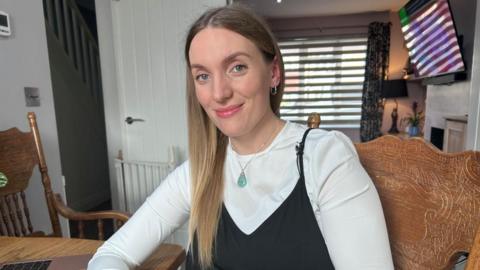 Alex McCarthy in her home, wearing a white top, black dress, a necklace, with a green stone is around her neck, she is smiling and looking at the camera, with long dark hair. She is sitting on a wooden chair, with another wooden chair to her right. There is a window, with blinds on it, behind her, and a white door, that is open.