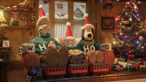 Wallace and Gromit sitting on a sofa with a gnome between them. They are all wearing santa hats and green winter jumpers. There is a Christmas tree next to them and a train set under their feet.