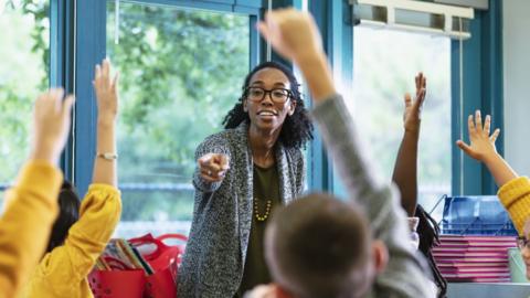 A teacher wearing black glasses and a grey cardigan smiles as she points towards a student with their hand in the air, surrounded by classmates also with their hands up.