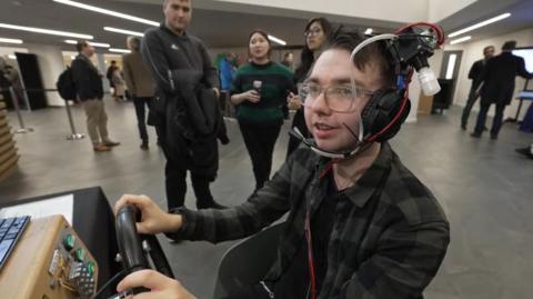 A man wearing glasses and a green check shirt smiles as he wear a set of headphones with wires and tubes attached to deliver smells in front of the user's nose. He has his hands on a steering wheel playing an arcade game.