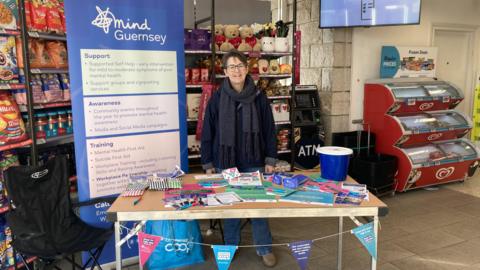 A woman in a navy jumper and grey scarf, stands behind a table with leaflets, pens and a donation bucket, in front of a blue sign which says Guernsey Mind.