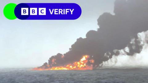 A grainy photo taken in the north sea off the east coast of England. It shows oil on the surface of the water that is on fire, with bright orange flames and a huge plume of dark grey smoke billowing into the sky. 