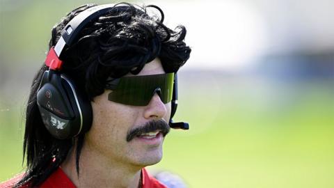 Content creator Dr Disrespect at a golf tournament, wearing a red tshirt and a black and red headset.