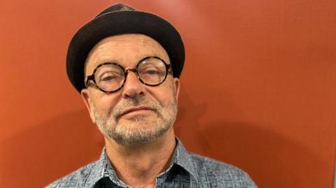 A man with a short grey beard is wearing a hat and looking at the camera. He has round brown glasses on and isn't smiling. He has a blue shirt and behind him the background is orange.