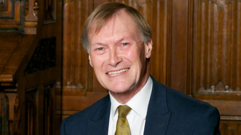 Sir David Amess, smiling, wearing a navy suit and olive green tie in front of wooden panels.