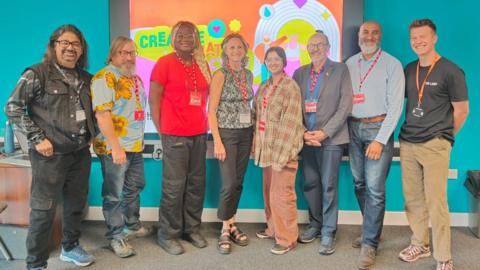 Eight people stand in a line. They are a mix of ages and background. There are three women in the centre and five men either side of them. There is a brightly multi-coloured background behind them.
