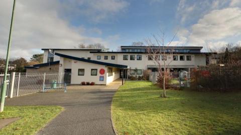 A school building which has a flat roof and is rendered in cream, with a path, grass and playground at the side.