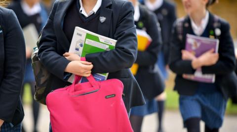 Pupils walking through a school playground, carrying books and bags. The school uniform is blue. You cannot see their faces. 