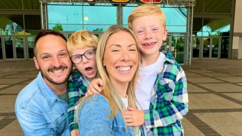 Daisy Woods, pictured second right, smiles in a selfie with her sons Sonny (left) and Oscar (right) and her husband Andy (far left). Both her and Andy are wearing denim shirts, while Sonny and Oscar are wearing matching white t-shirts and green, blue and yellow checked shirts over the the top.