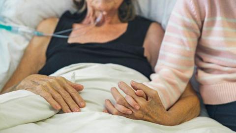 A woman with tubes coming out of her nose lays on a bed and holds the hand of a child. Their faces are not visible.