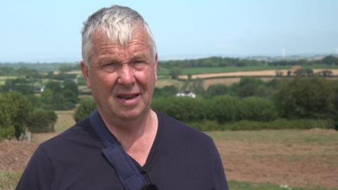 John Gibson is wearing a blue top and is standing in front of a field with a green hedge in the background. It is a sunny day.