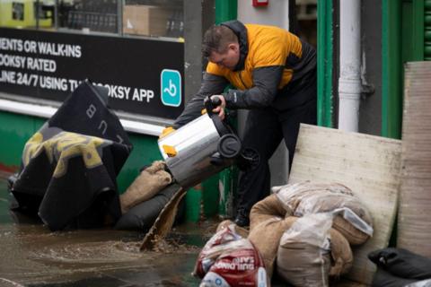 Business owner stood in shop opening, poruing out a binfull of water into a flooded street which is full of muddy water, sandbags and rubbish