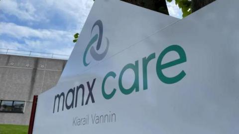 A white sign, with a peak, that reads Manx Care in English and Manx in grey and green writing below the Manx Care logo.