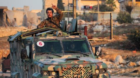 A man in a camouflage jacket stands in the back of a van driving down a road, with one hand in the air.