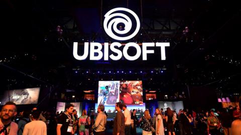 An illuminated Ubisoft sign hanging from the ceiling of a warehouse where a games convention is taking place