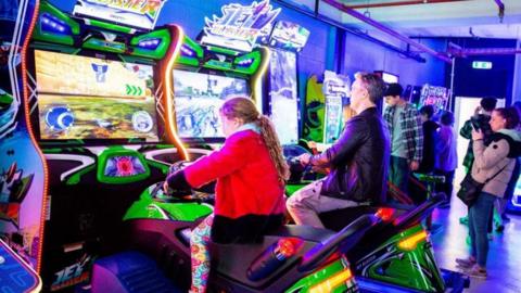 People are playing arcade video games. Two people, a young girl and a man, are playing games by sitting on futuristic motorbike-type vehicles.