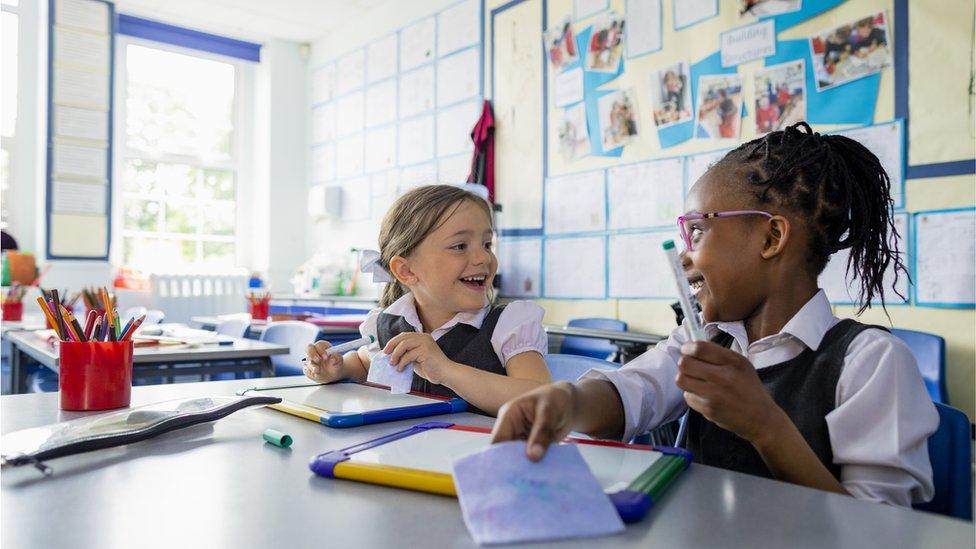 Two children smile at each other while sat at their desks in a primary classroom.
