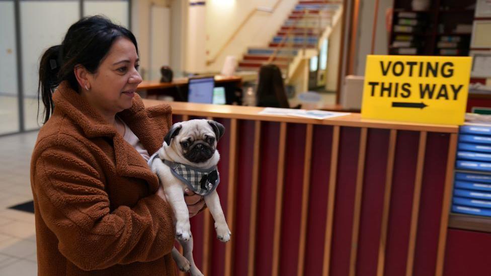 A woman holds a pug as she passes a yellow 'Voting this way' sign