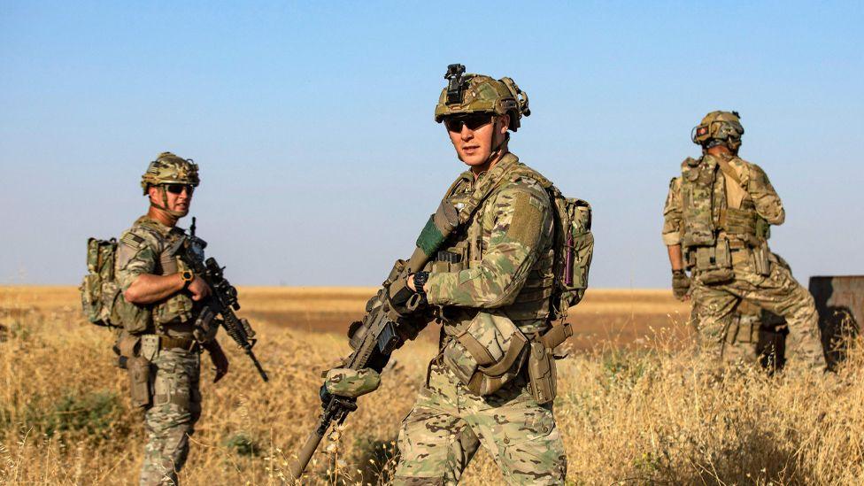 A group of three US soldiers wearing camouflage and carrying rifles patrolling in a field near the Turkish border in Syria