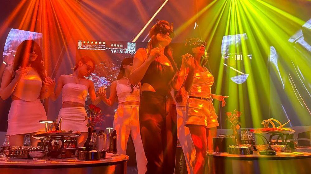 Seven women in a karaoke bar, wearing white, black and gold clothing, holding hands and dancing in neon lights