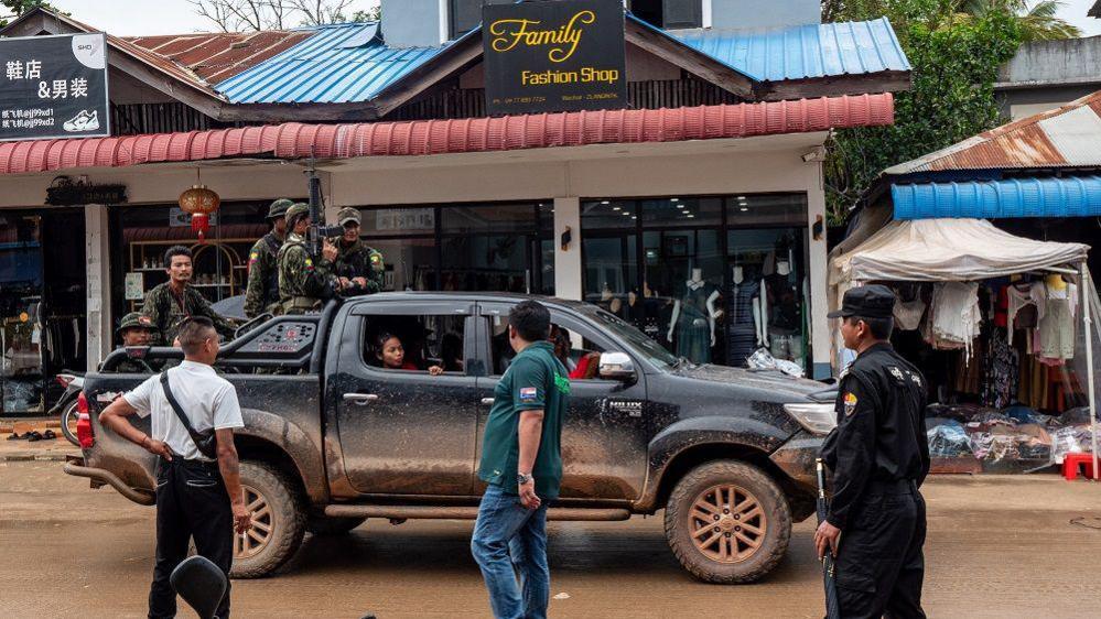 A vehicle inside the city. Armed men who work for the local warlord in fatigues stand on the back of a black pickup truck driving down a road with shops on it, one called Family Fashion Shop