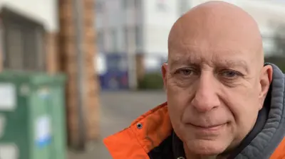 Dave wears a bright orange and grey work anorak and a grey hoodie. He is bald and stands next to the road outside the Vauxhall factory. The Vauxhall emblem is on one corner, and the building recedes into the distance with many windows across its two storeys.. 