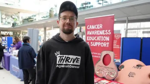 Shaun Whitmore/BBC Toby Freeman, standing by his charity stall within The Forum in Norwich. He is wearing a black hoodie with a logo stating "Thrive against cancer" and wearing a black baseball cap. He has a full beard and moustache and wears glasses.