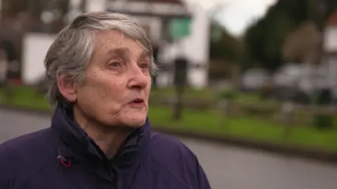 Justine, a woman in her 60s with short white hair and wearing a dark blue waterproof jacket, is standing by a road. There are buildings on the other side of the road in the background. 