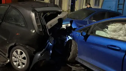 A black car and two blue cars with severe damage to the rear of the black car and the front of the blue car.