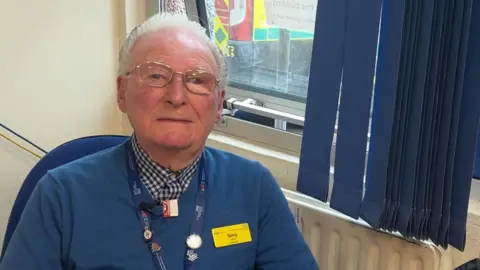 Terry Seston, who has grey hair and gold glasses is wearing a checked shirt and blue jumper, sits in front of a window. Behind him, out of the window, can be seen a yellow and blue ambulance vehicle.