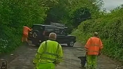 A screenshot of dashcam footage showing a 4x4 reversing into a roadworker wearing orange hi-vis clothing.