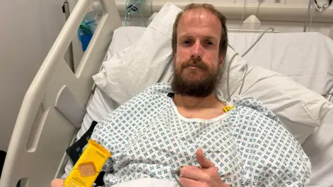 Nathaniel Dye in a hospital gown in a hospital bed. He gives the camera a thumbs up symbol.