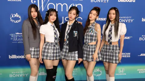 The five members of NewJeans stand against a blue and turquoise backdrop, dressed in variations of tartan school skirt with blazers and ties. 