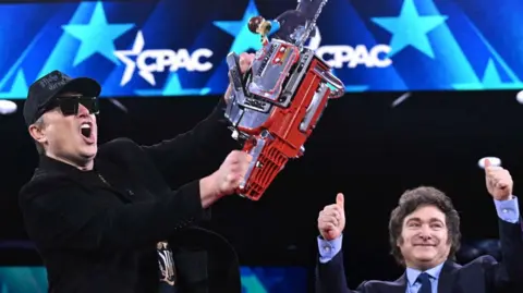 Elon Musk holding chainsaw on stage next to Javier Milei holding two thumbs up