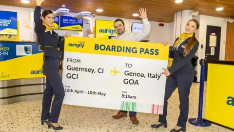 Three Aurigny staff are holding a giant boarding pass in the airport. The ticket said FROM GUERNSEY CI TO GENOA ITALY, Date 12th April - 10th May, Time 8.15am. The two women are wearing navy trouser suits with yellow detail and the man is wearing a white shirt, jeans and brown shoes. There are Aurigny yellow and blue banners and posters in the background.