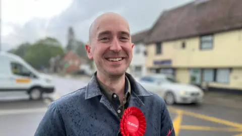 Ben Goldsborough smiling whilst wearing a grey jacket over a green shirt. He has a red Labour rosette on and is standing by a junction with traffic behind him.