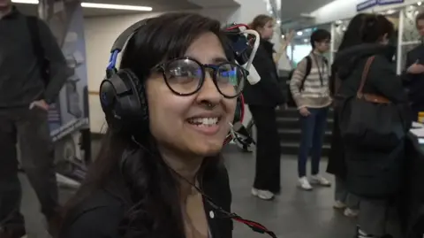 A woman wearing glasses with long dark hair smiles while looking off the camera. She's wearing the experimental headset of headphones with a plastic tube attached to deliver smells from a bottle to the user's nose.