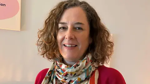 Lisa Gibson is smiling at the camera while standing indoors. She has brown curly hair that falls just above her shoulders. She is wearing a patterned scarf and pink top.