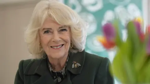 Queen Camilla in a dark coat with a bee broach and some flowers in the foreground.