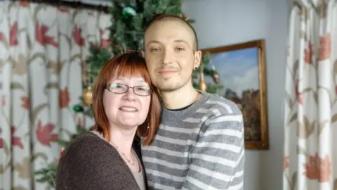 Supplied Robin Freeman, who was diagnosed with testicular cancer in his early 20s, has short hair and is wearing a grey and white striped top. He is hugging his mum, who has auburn hair and is wearing a brown jersey. They are pictured in a room which features floral patterned curtains and pictures in the background.