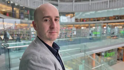 Paul Moseley/BBC Ben Goldsborough is looking at the camera over his right shoulder. He has a grey suit jacket on over a blue shirt. He is standing on a glass internal balcony, with the library visible behind him.