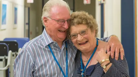 Shrewsbury and Telford Hospital NHS Trust Terry Seston, a man with grey hair and glasses is wearing a blue shirt, with navy and red stripes. He has his arm around his wife Babs, who has curly blonde hair and glasses and is wearing a blue polka dot top. Both are smiling, and Babs is holding Terry's hand, which lies on her shoulder. Behind them can be seen a hospital corridor, with blue wheelchairs along the walls to their left hand side.