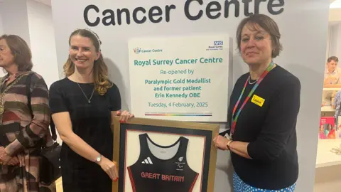 Two women stood in front of a plaque which reads 'Royal Surrey Cancer Centre re-opened by Paralympic golf medallist and former patient Erin Kennedy OBE Tuesday 4 February 2025. 