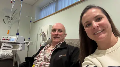 Supplied Angharad Binder, with long straight hair, wearing a white top with black stripes. She is next to her father Robert Bendall, who is undergoing treatment for bowel cancer. He is wearing a checked shirt and black jacket, and he has close cropped hair.