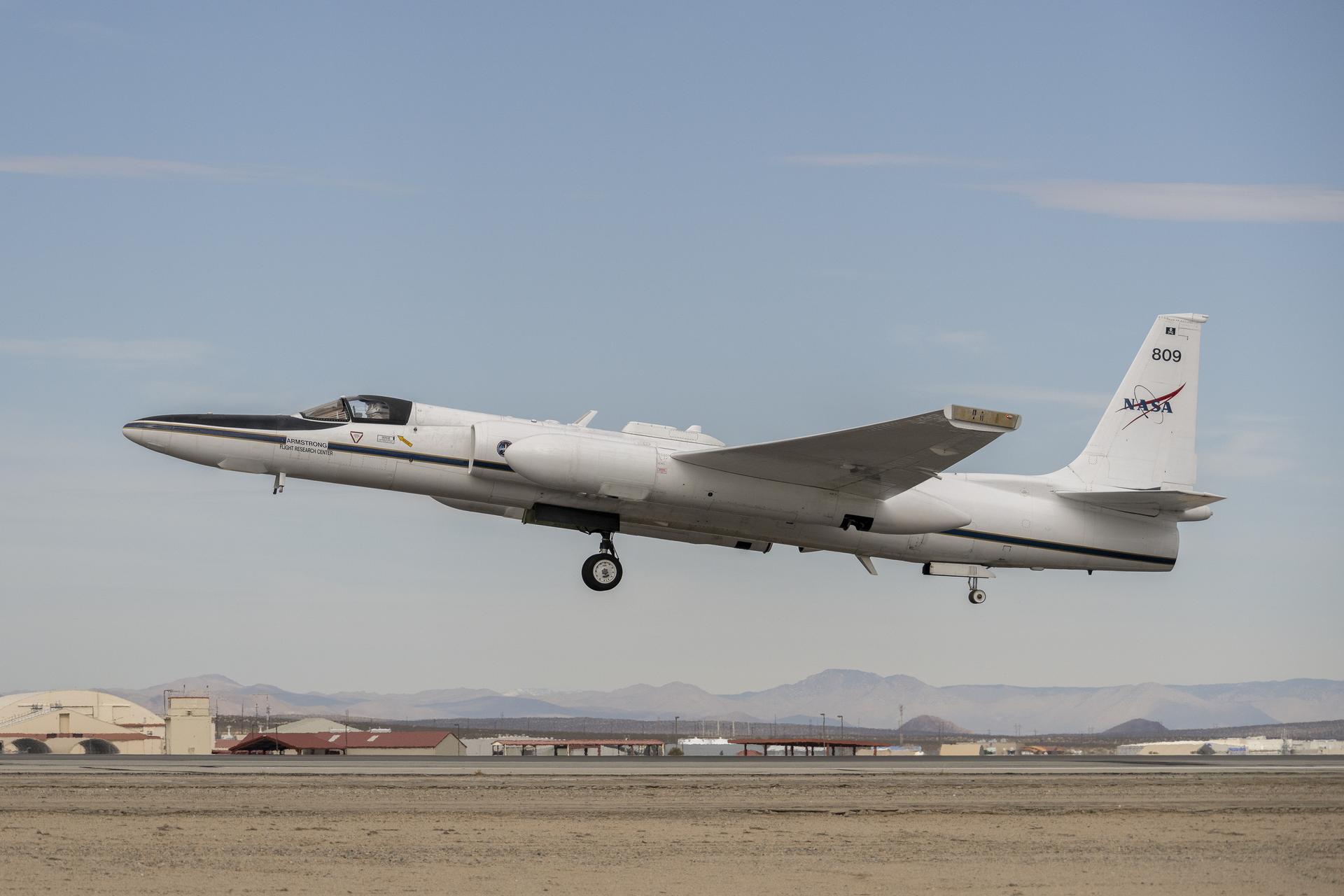 A plane begins to ascend from the runway on dim desert morning, heading toward the left of the frame. The wheels of the plane are still dropped and the nose of the plane is pointed upward toward higher skies, its tail featuring the NASA insignia. The helmeted pilot inside is barely visible through the cockpit window. Below a desert landscape with indiscernible buildings are flanked by a hazy mountain range in the distance.