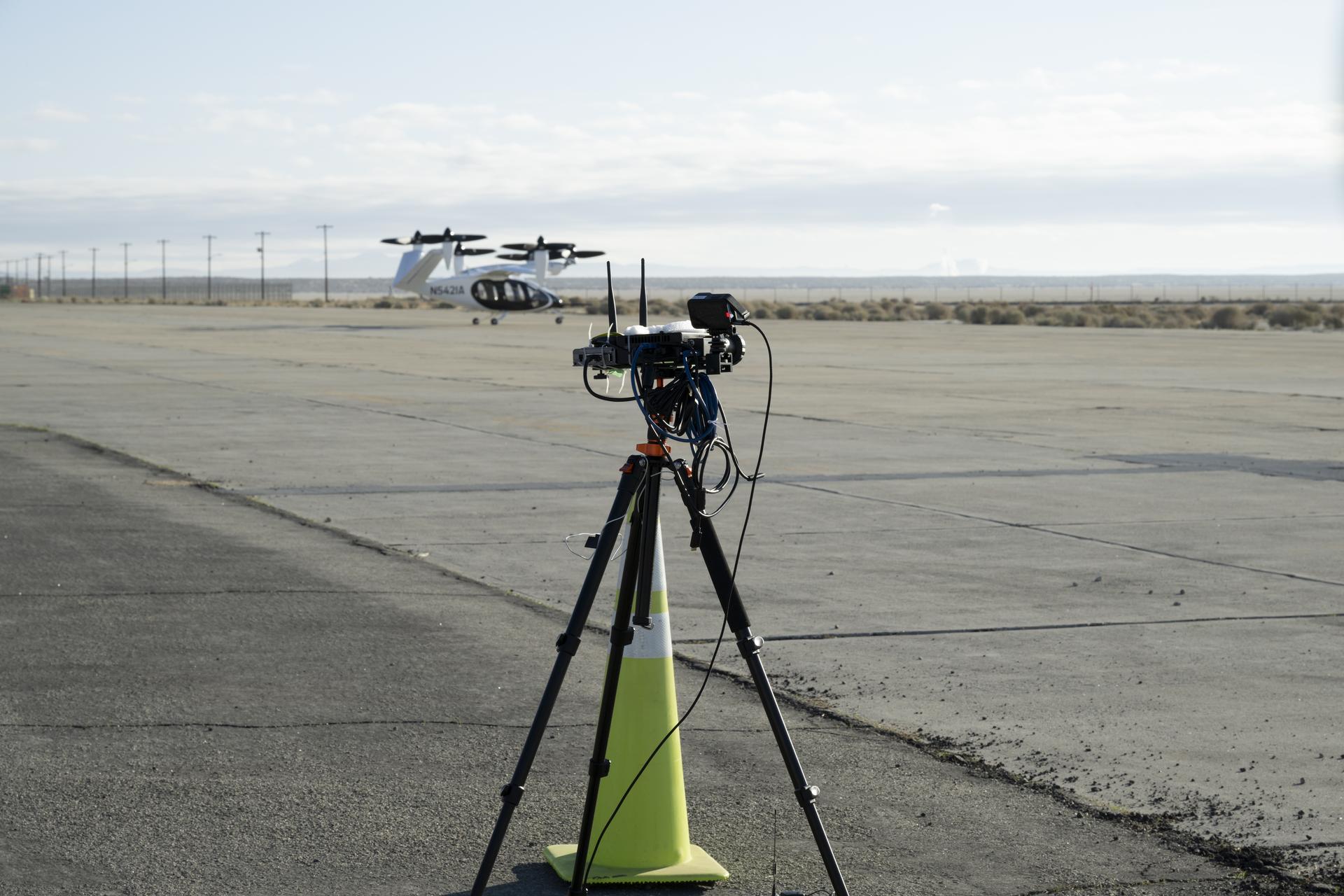 A yellow traffic cone and a black tripod with black testing instruments stands in the foreground on a concrete pad with a desert landscape, power lines, and a black and white aircraft in the background. The aircraft has six black propellors that sit on white arms and connect to the aircraft body, which has black doors and is pod-shaped. The aircraft sits on three small wheels.